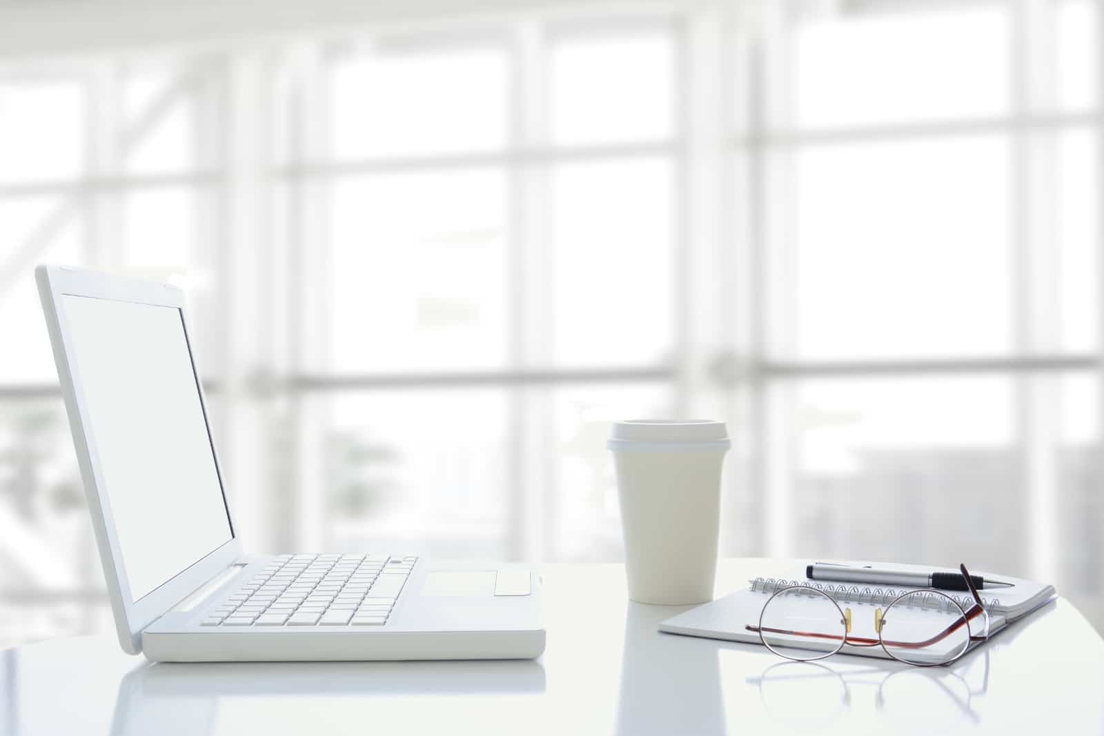 laptop on a desk with cup, notebook, glasses in front of windows